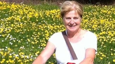 Danielle smiles at the camera in a grainy photograph. She has blonde hair and wears a white t-shirt. She is sitting in a meadow or garden with long grass and lots of daisies and buttercups.
