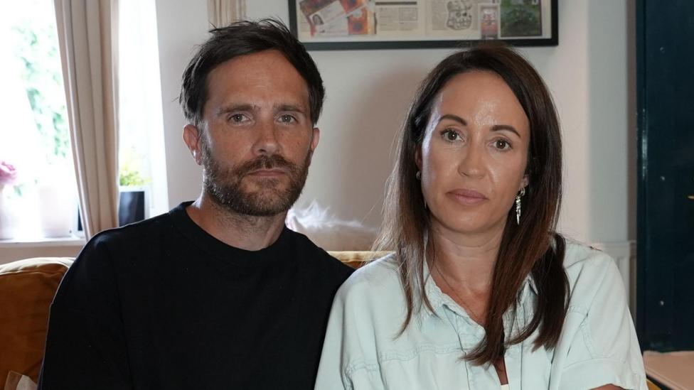 Craig and Carly Bowe sitting on a sofa. Craig is wearing a black T-shirt. Carly is wearing a turquoise shirt and has long brown hair.