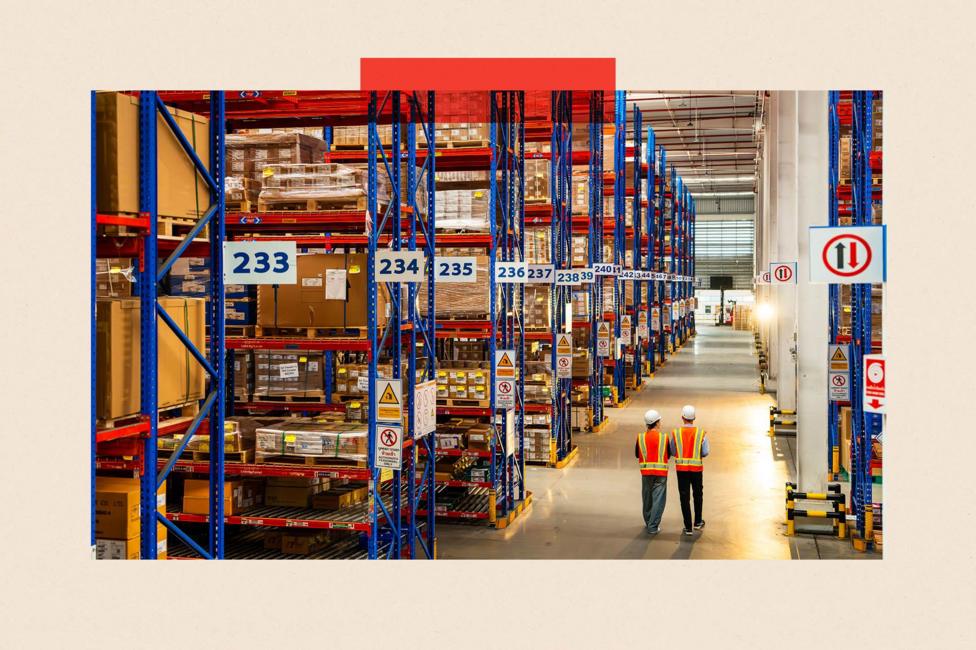 Two people in white hats and orange vests walk across a warehouse