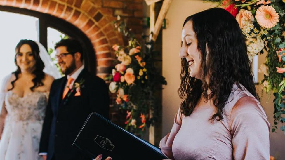 Zadie Ward, in a pink dress, reads from a black folder at a wedding