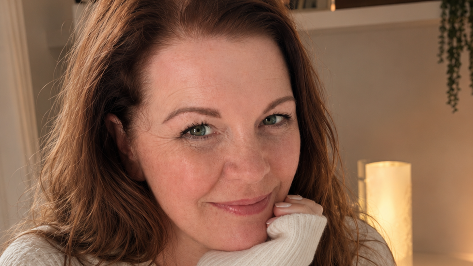 Samantha Thomas is pictured with long auburn hair wearing a cream knitted jumper, resting her chin on one hand and looking towards the camera. Warm indoor lighting, framed photographs and houseplants are visible on shelves in the softly blurred background.