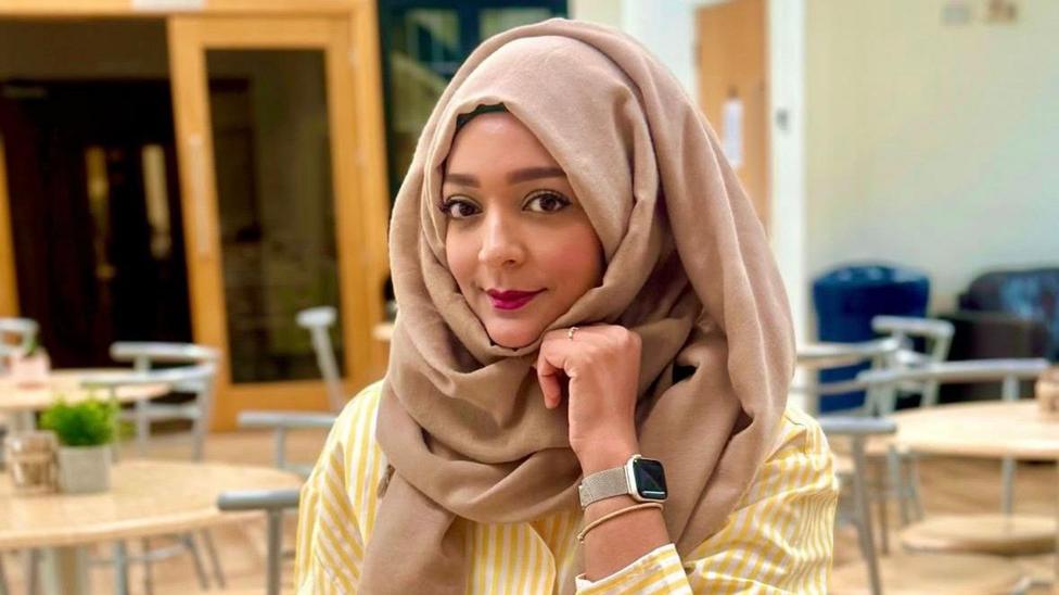 A woman wearing a brown hijab and yellow and white striped top smiling, she has her chin resting on her hand and she has a watch on her wrist.