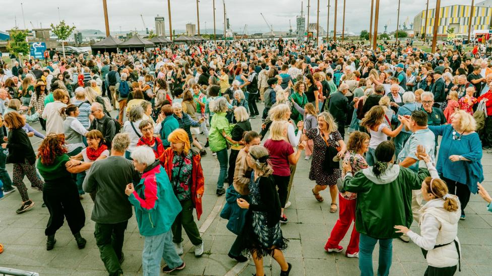 A very large crowd of people are dancing. Some trees, poles and buioldings are in the background.