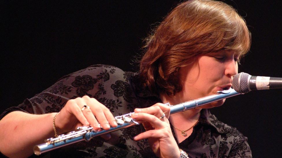 A woman is onstage playing a flute. She has short brown hair and is wearing a watch, a bracelet and rings on both hands.