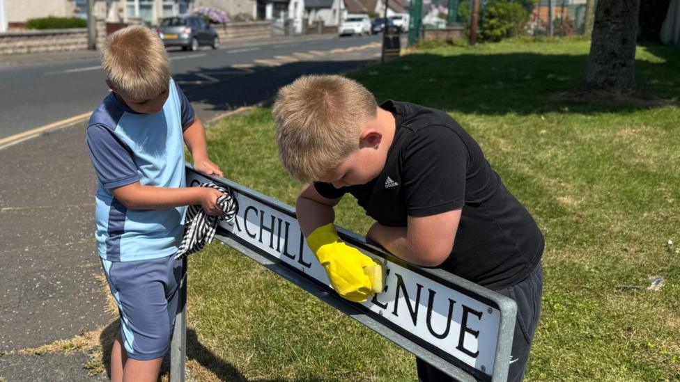 Two children are cleaning a street sign that reads