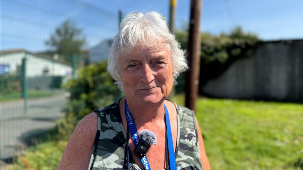 The image shows an older woman standing outdoors on a sunny day, with a clear blue sky behind her. She has short white hair and wears a sleeveless camouflage-patterned top. Around her neck hangs a blue lanyard that reads Primary School, along with a small microphone covered by a windscreen, suggesting she might be giving an interview or recording something.