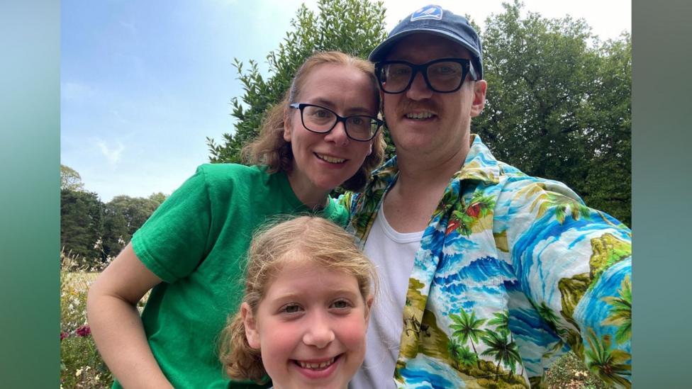 A family of a man, woman and their child. The man is wearing a hawaiian shirt and a blue cap, and is smiling at the camera