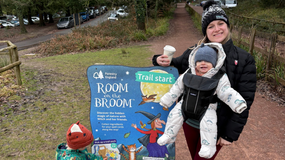 A woman wearing a black coat and bobble hat, with a babyin a sling on her front, standing next to a forestry england trail sign