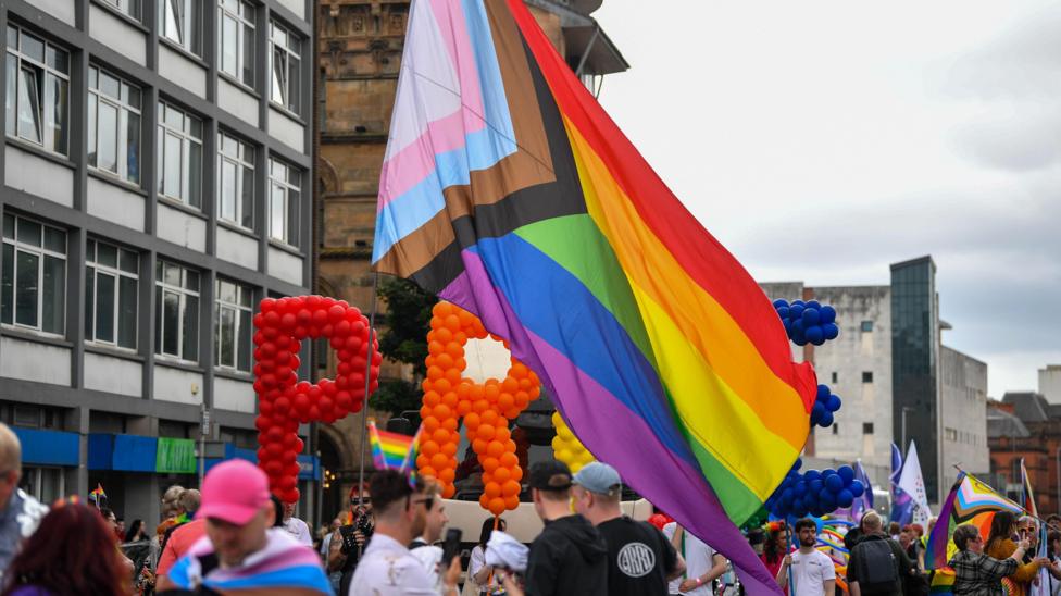 A large pride flag being flown above a crowd of people.