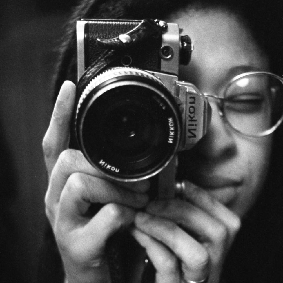 A close-up of Sandra, a young black woman with metal-framed glasses, holding a Nikon camera on its side and looking through the view-finder.