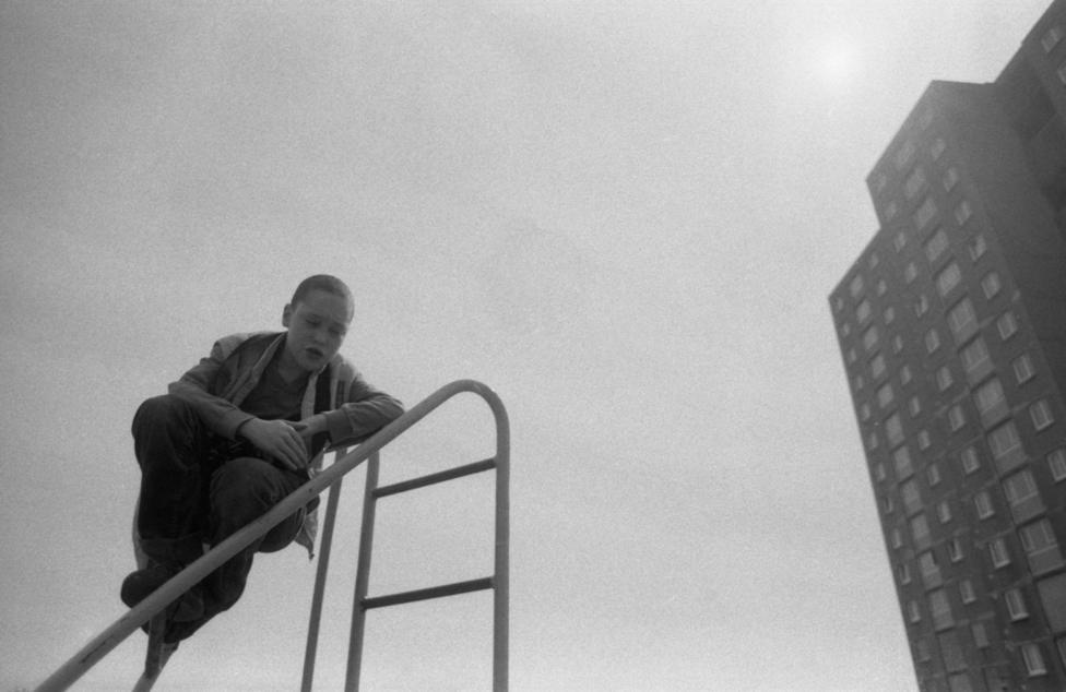 A boy in dark trousers and a light jacket sits on a climbing frame in front of the high flats of Greendykes. The sun is seen above the building.