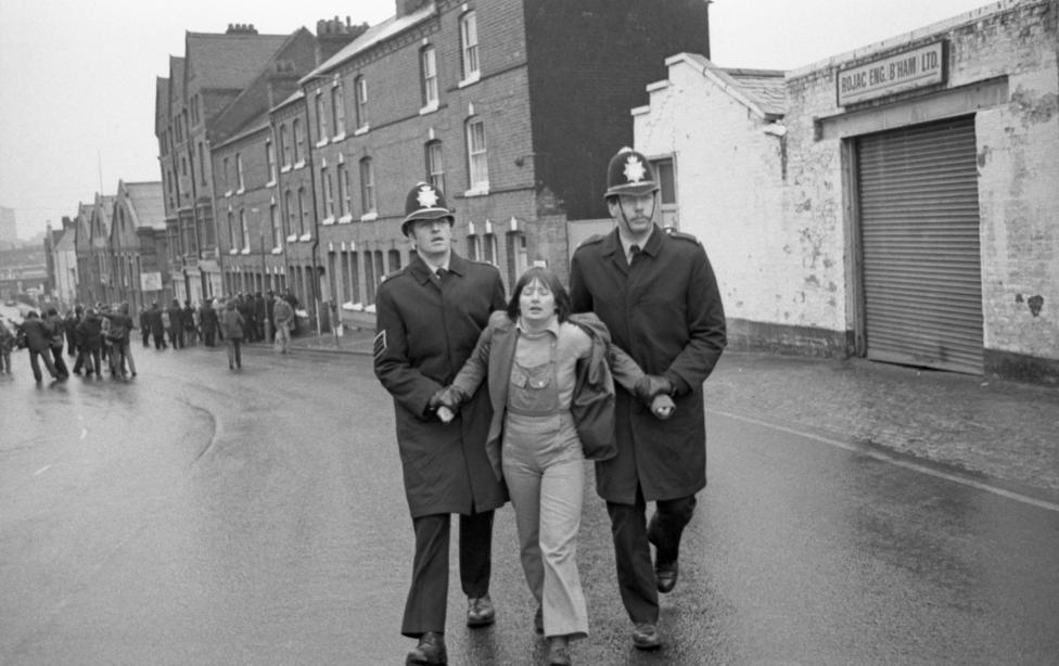 A young woman in dungarees in 1981 is being marched by two police officers in long coats and helmets up a street in front of a garage. A crowd can be seen in the background.