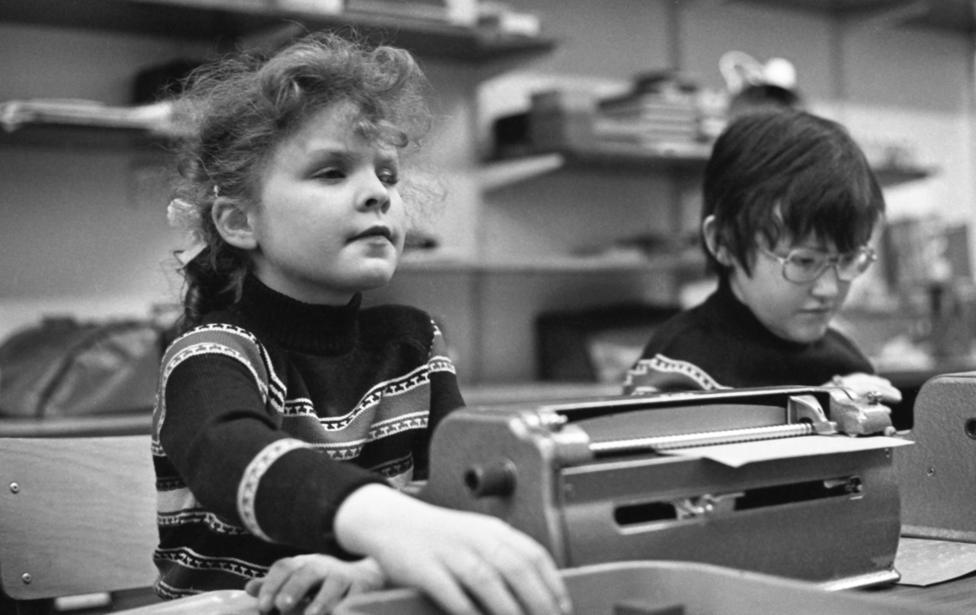 Two blind children working brail machines in a room. They are wearing identical jumpers and are concentrating on what they are doing.