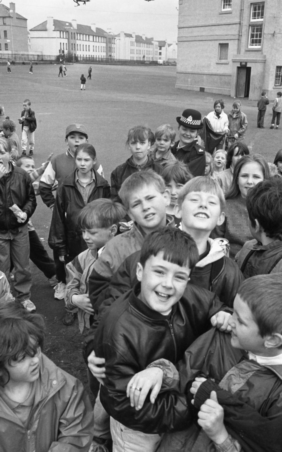 A group of about 20 children crowd into frame in front of a school. They are laughing and enjoying themselves.