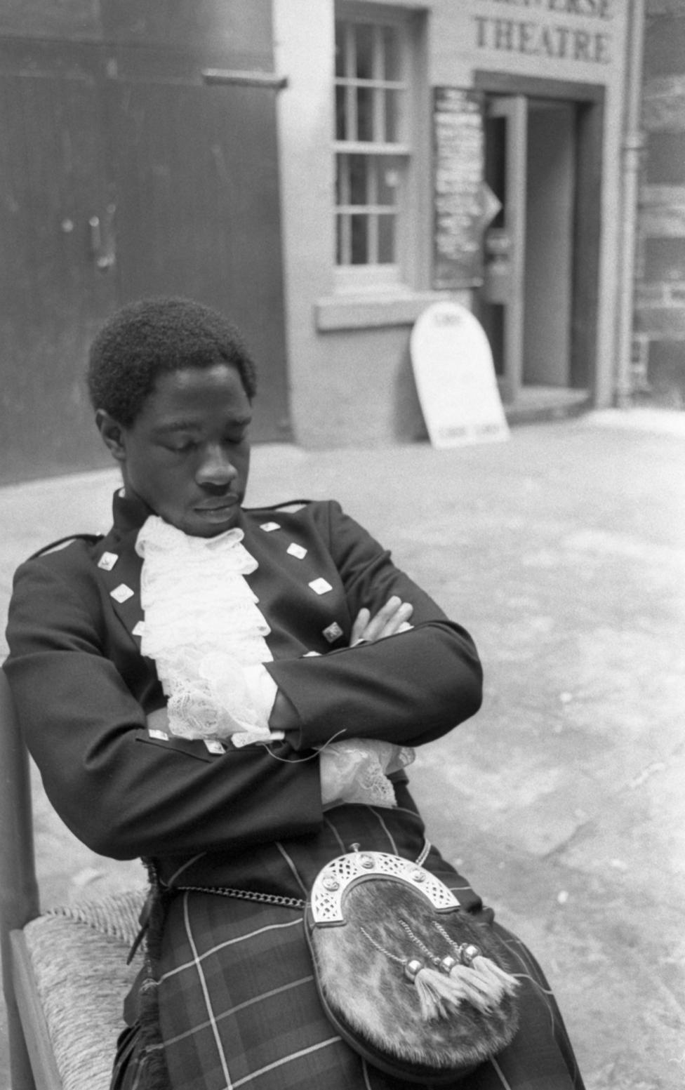 A young male member of a black theatre group in Craigmillar in 1983. He is sitting apparently sleeping in front of a shop, in full tartan dress.