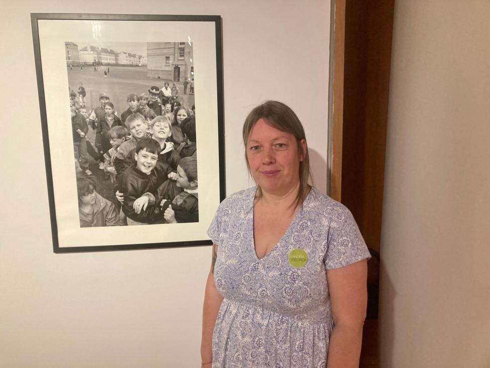 Becky Smith is in a grey patterned dress. She has long blonde hair and is standing in front of a black and white photo.
