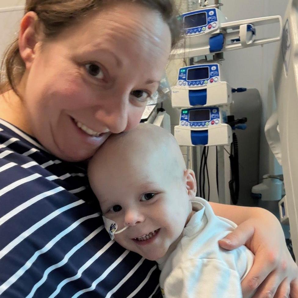 A woman with tied-back brown hair wearing a navy and white striped top. She is hugging a young boy wearing a white t shirt, who has a feeding tube in his nose. They are sat with hospital equipment behind them and are both looking at the camera and smiling.