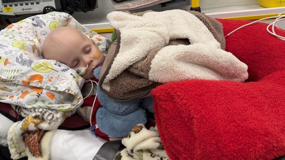 A young boy with a feeding tube in his nose lying down with his eyes closed. He has lots of different coloured blankets piled on top of him.