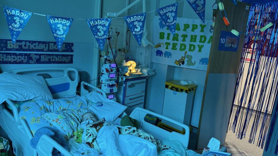 A young biy in a hospital bed, wrapped in cartoon-print blankets. Above him is blue bunting which says