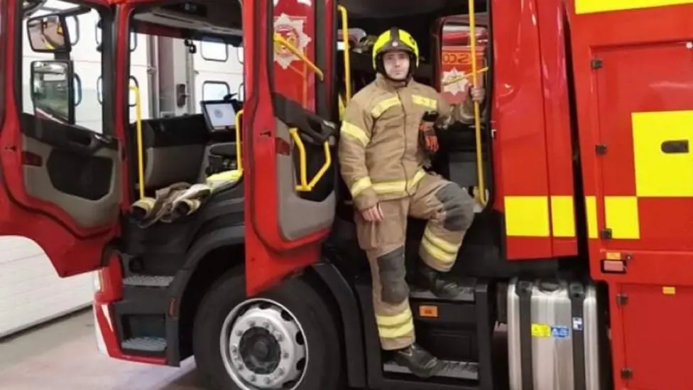Barry Martin is standing on the steps of a red fire engine, looking at the camera. He is wearing brown fire-resistant overalls with hi-vis stripes on them and a yellow and black helmet