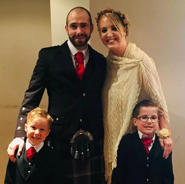 Barry an Shelley Martin with their twin boys Oliver and Daniel who look to be aged about five in the photo. They are all dressed up as if going to a wedding. Barry and his sons are wearing kilts and formal Scottish attire and Shelley is wearinga cream shawl and flowers in her hair