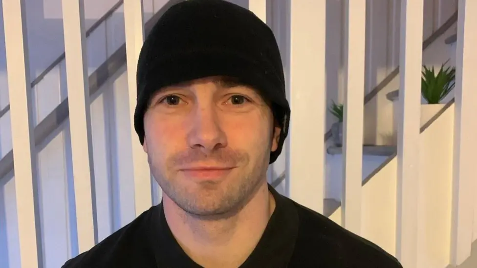 Barry Martin is smiling at the camera. He is wearing a black top and a black beanie hat and is standing in front of stairs in a house