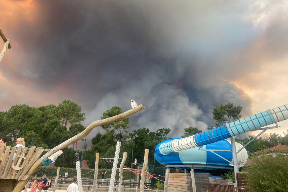 A huge cloud of dark grey smoke approaches a campsite where children's play equipment can be seen in the foreground.