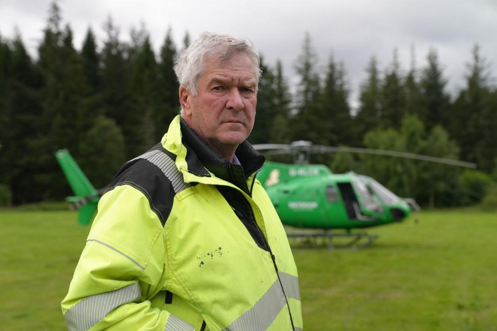 Nethy Bridge community council leader Sandy McCook wearing a high-visibility yellow jacket stands in a grassy clearing with a green helicopter parked behind them. Dense conifer forest forms the background under an overcast sky.