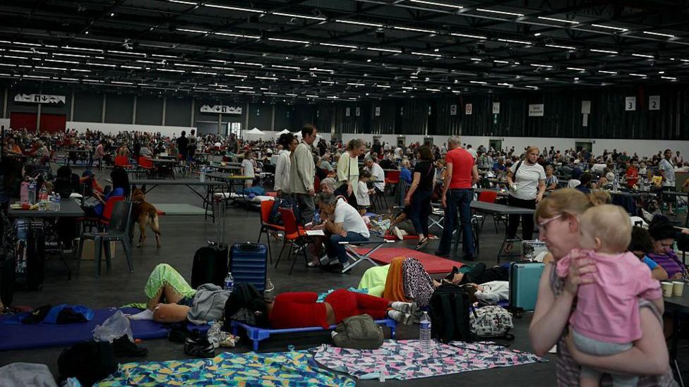 A number of people shelter at an exhibition centre - some people sleep on mats on the floor, while a woman carries a baby on the bottom right of the photo