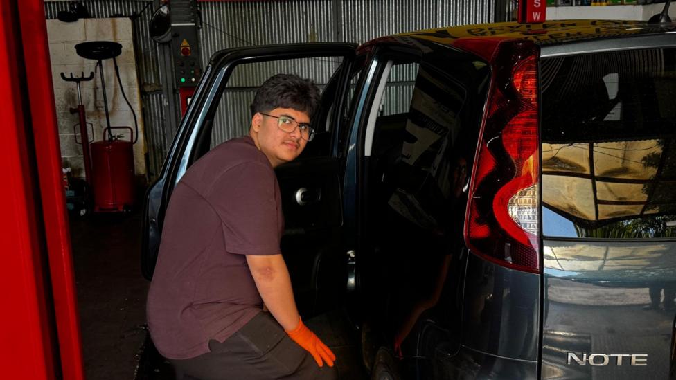 Azhe is seen wearing orange gloves as works on a dark-coloured car inside a vehicle workshop. The rear door is open, with garage equipment and automotive service banners visible in the background.