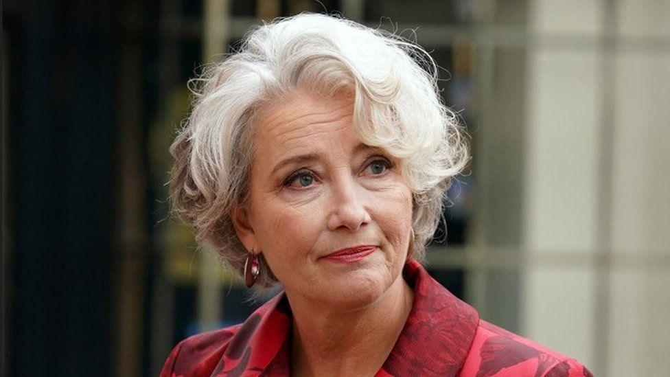 Close up of Emma Thompson from the shoulders up, who has short, white hair and is wearing a red patterned jacket.