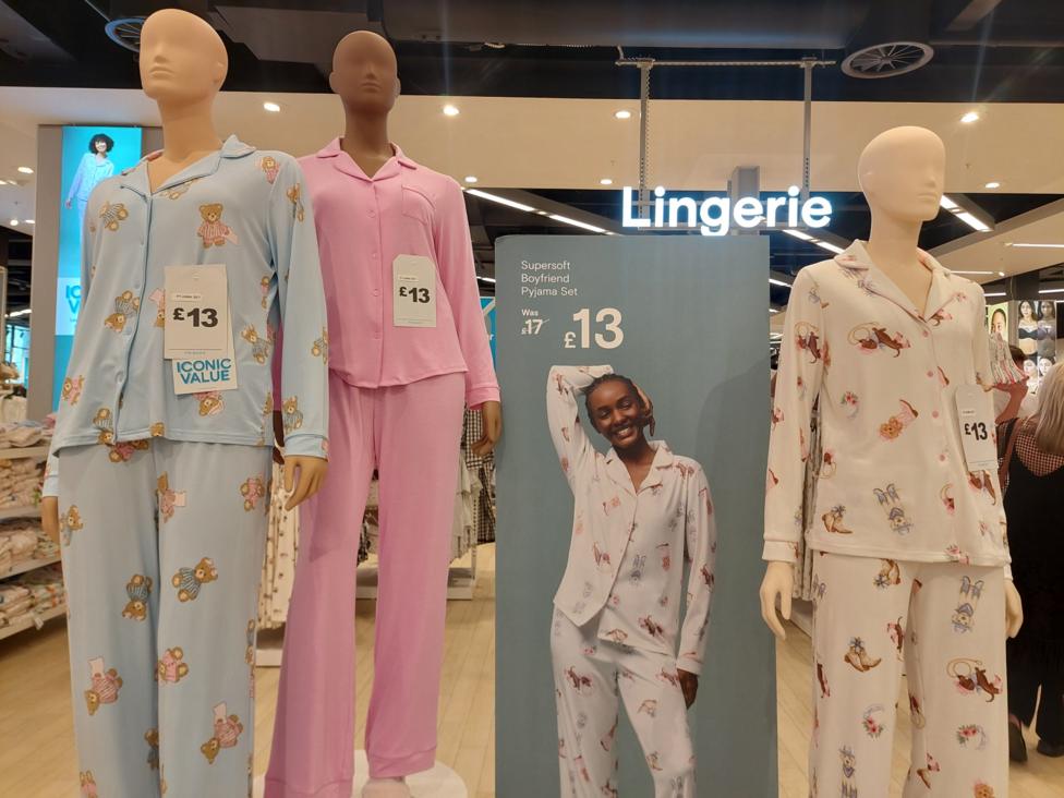 Three mannequins wearing long-sleeved pyjama sets stand in the foreground of a lingerie section. One mannequin wears a light blue set with teddy bear prints, one wears a plain pink set, and one wears a white set with illustrated character prints. Price tags on the pyjamas show £13. Between the mannequins, a promotional sign advertises a “Supersoft Boyfriend Pyjama Set” reduced from £17 to £13. An illuminated “Lingerie” sign hangs above, with shelves and clothing displays visible in the background.