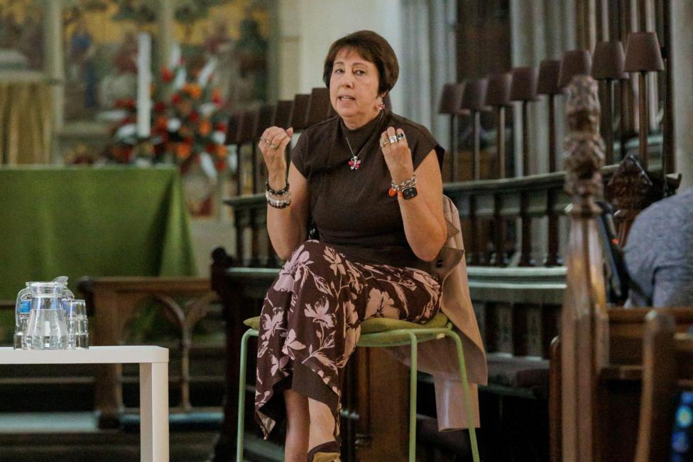 A woman is sat at the front of a church speaking to an unseen audience. She has a brown top on and brown-patterned skirt.