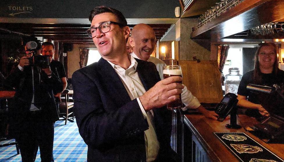 A man with square glasses and short dark hair laughs and turns away from a bar holding a pint of beer. He is wearing a white shirt and dark jacket. There is a woman behind the bar laughing and a bald man is also pictured behind the man holding the beer laughing. Another man is holding a television camera in the background.