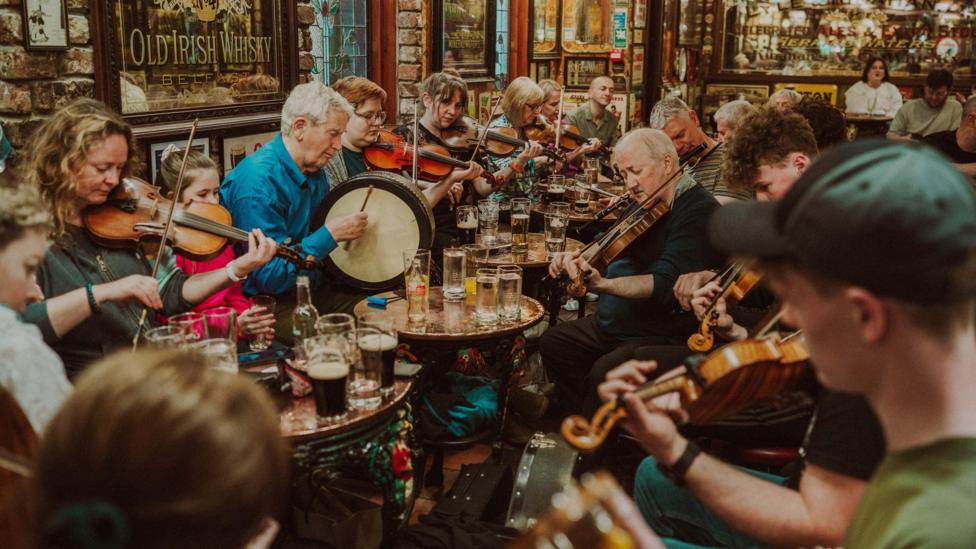 People sitting in a pub are playing instruments. Many are playing the fiddle. The pub is crowded. Drinks are sitting on tables.