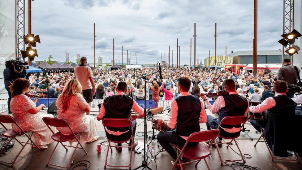 A photo taken from behind a band on stage. The band is sitting and holding instruments including the fiddle and drums. A huge crowd is dancing. Some have their arms in the air. It's cloudy.