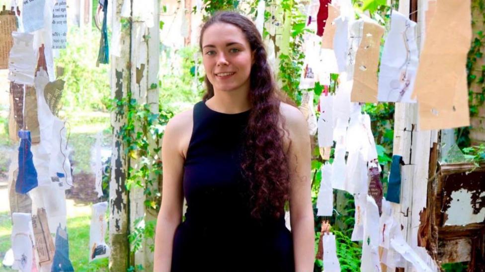 Eimear is standing. She's smiling and looking into the camera. She has dark purple wavy hair and is wearing a dark blue dress. There are pieces of paper hanging up around her. Leaves are behind her.