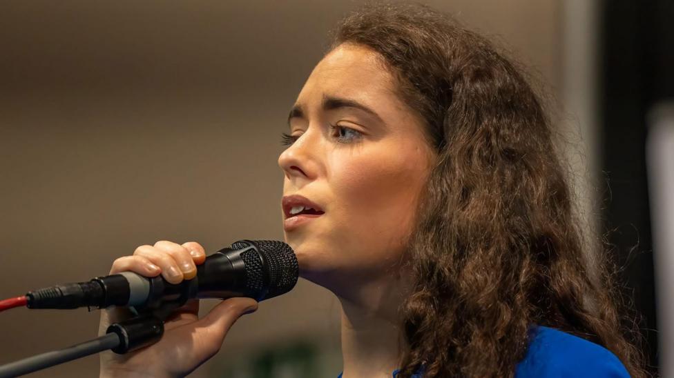 Eimear is onstage singing. Her brown hair is wavy. She's holding a mic and is wearing blue.