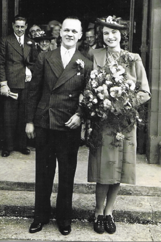 Stan and Beatrice pose in a black and white photo. Stan is wearing a pinstripe suit with a button hole, Beatrice holds a large bouquet of orchids and is wearing a period dress and jacket with dark hat and shoes