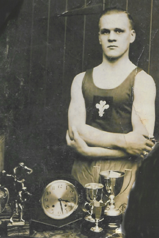 Stan, in his 20s, poses defiantly before the camera with arms crossed. His vest bears the three feathers Welsh emblem. In the foreground can be seen several silver boxing trophies and an ornamental clock