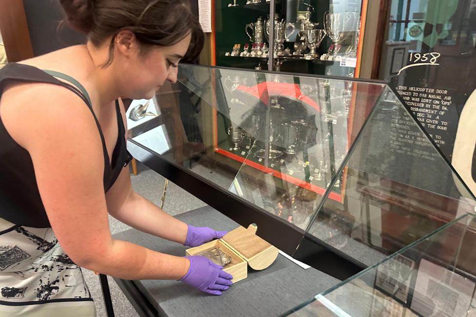 Wearing protective purple rubber gloves, the Curator, wearing a cream and black skirt and black sleeveless top, uses a red rubber sucker to open a hinged display case in order to avoid transferring fingerprints onto the glass.
