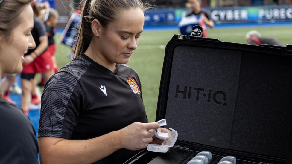 Ffion holding a white mouthguard in her hand. She stands next to a black case with multiple mouthguards in. She wears a black rugby t-shirt and stands on a rugby pitch.