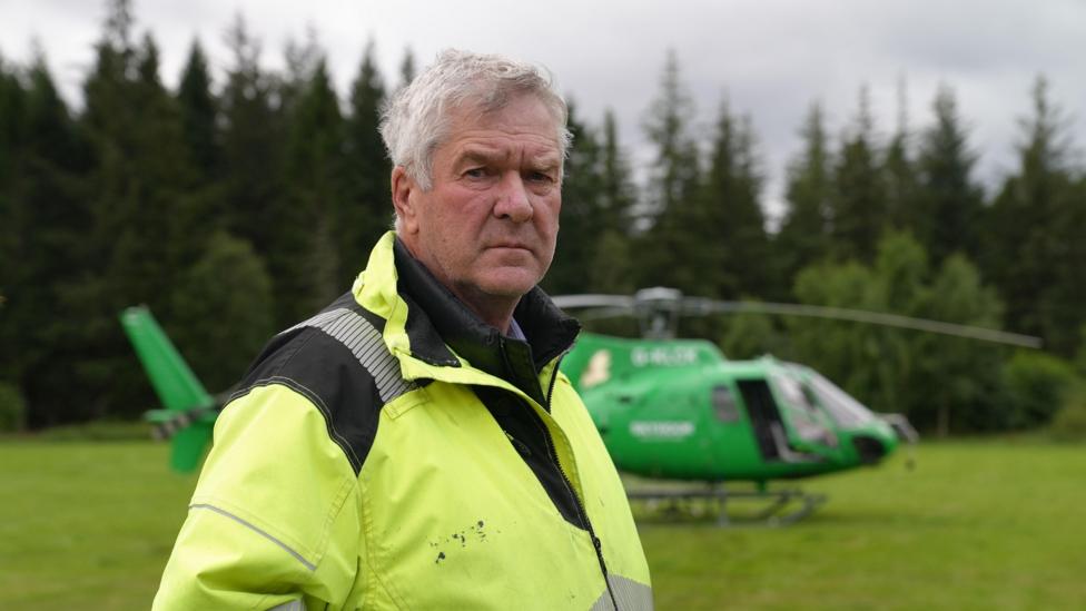 Nethy Bridge community council leader Sandy McCook wearing a high-visibility yellow jacket stands in a grassy clearing with a green helicopter parked behind them. Dense conifer forest forms the background under an overcast sky.