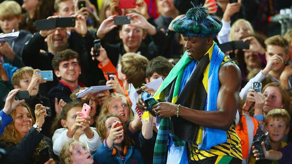 Usian Bolt signs autographs for fans waiting trackside during the Commonwealth Games in 2014. Bolt is wearing a tartan hat, and has a Scottish flag and tartan scarf wrapped around his shoulders. He is wearing his running gear in yellow and green colours. The fans are all smiling and reaching out with things for him to sign.