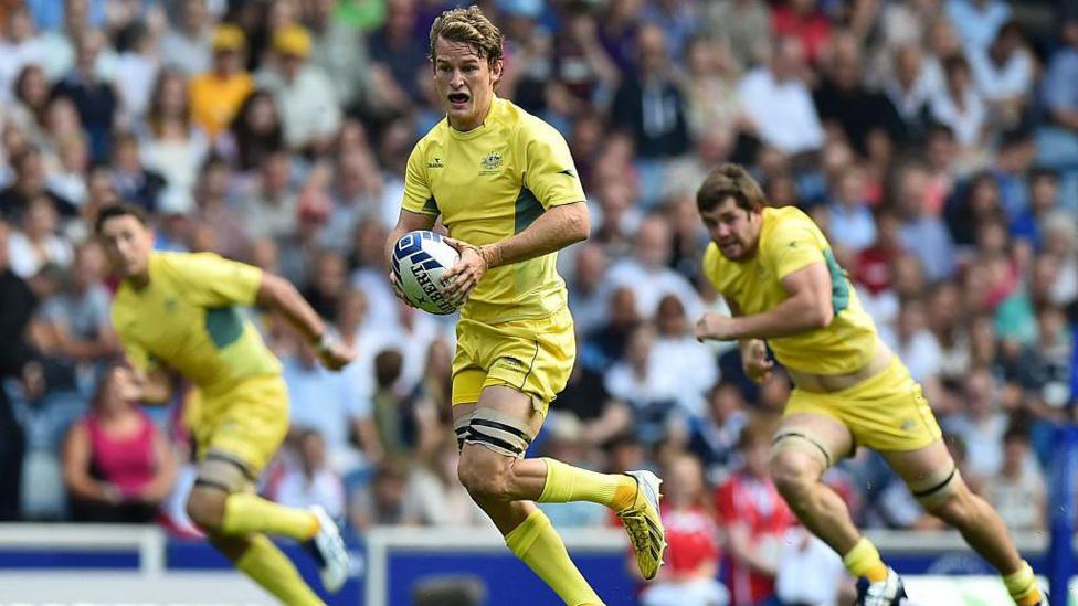 An Australian rugby sevens player runs forward holding the ball, with two team mates behind him. They are wearing a strip that is nearly all yellow, with a little green on the jersey.