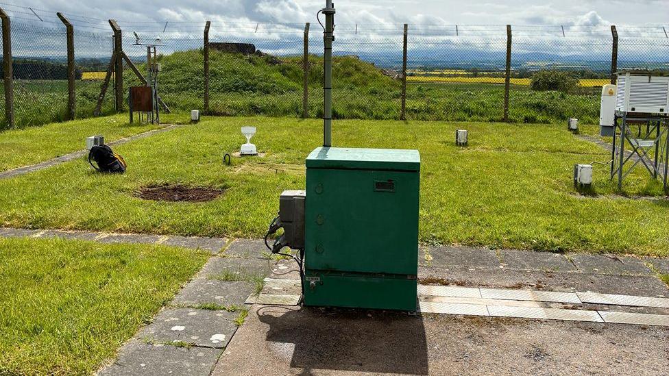 A weather station with a large green box in the middle of it and fencing round the outside