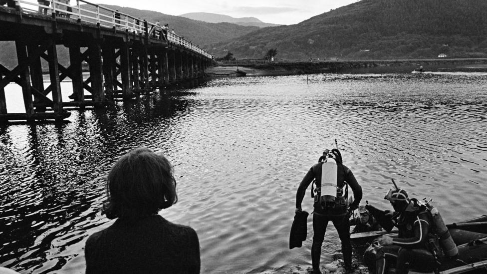 Black and white image looking across a stretch of water, with a large wooden bridge on the left crossing an estuary. In the foreground, a woman with her back to the camera is looking out, as two divers in scuba gear prepare to enter the water. One is standing with flippers in his hand and a white air tank on his back - the other is sat in a boat on the water's edge