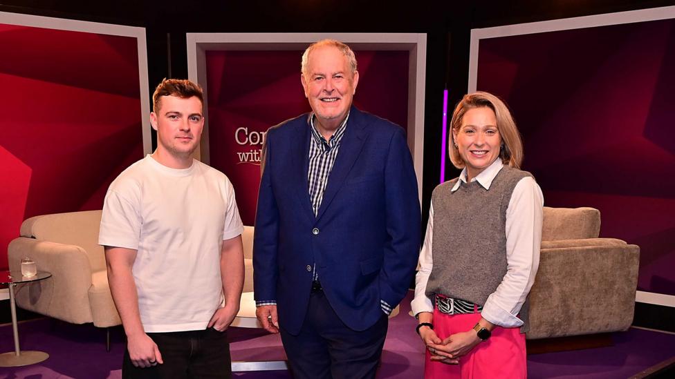 Three people standing beside each other, two men and a woman. The man has short dark hair and is wearing a white t-shirt. The man has short grey hair and is wearing a blue blazer and checked shirt. The woman has shoulder length blonde hair and is wearing pink trouser, white shirt and brown jumper.