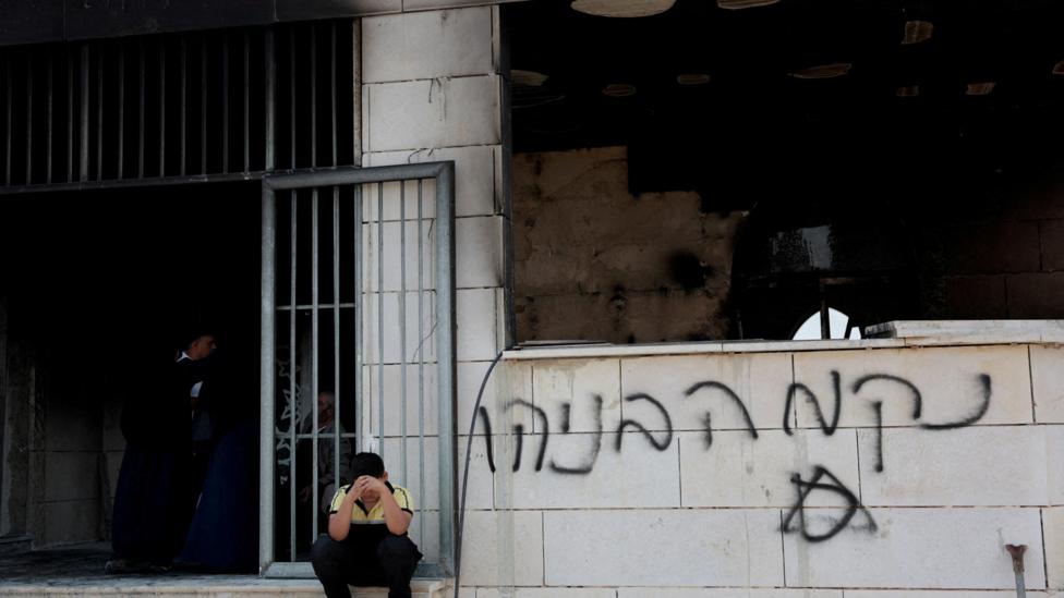 A boy sits with his face in his hands outside a damaged mosque which has graffiti on its walls