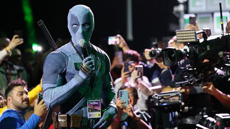 Ryan Reynolds, disguised in a Deadpool costume, holds a microphone and is surrounded by cameras and fans as he speaks at San Diego Comic Con.